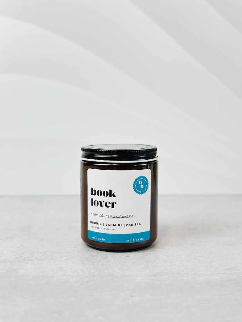 May include: A brown glass candle jar with a black lid. The label reads "book lover" and "orchid | jasmine | vanilla scented soy candle".