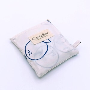 Peut inclure: Sac de courses réutilisable plié, de couleur crème, avec un motif de fruits d'agrumes bleus. Le sac porte une petite étiquette crème avec l'inscription "Cut & Sew Handmade in Canada". Une petite boucle crème est attachée en haut.