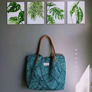 May include: A teal tote bag with a tropical leaf pattern hangs below four framed botanical prints. The tote has brown leather handles and a label that reads "Cut & Sew". The prints feature detailed watercolour paintings of green leaves.