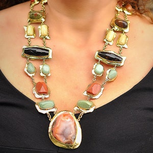 May include: A gold-toned statement necklace featuring a variety of colorful gemstones. The necklace has a layered design with rectangular, oval, and irregular-shaped stones in shades of brown, black, green, and red. The necklace is worn against a black top.