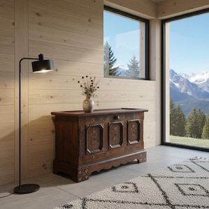 Könnte beinhalten: Eine dunkelbraune Holztruhe mit kunstvollen Schnitzereien und einem dekorativen Sockel. Eine kleine Vase mit Trockenblumen steht obenauf. Eine schwarze Stehlampe und ein gemusterter Teppich befinden sich im Vordergrund. Große Fenster mit Bergblick.