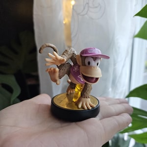 Könnte beinhalten: Eine braune und beige Figur eines Affen mit rosa Kappe und lila Weste, die auf einer goldfarbenen Basis steht. Die Figur wird in der Hand einer Person gehalten, mit einem Fenster und einer Pflanze im Hintergrund.