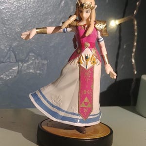 May include: A figurine of a character from The Legend of Zelda series. The figure is wearing a pink and white dress with gold accents. The figure is standing on a black base.
