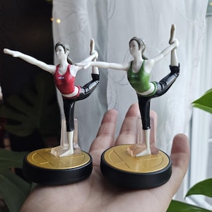 Puede incluir: Dos figuras de una mujer en postura de yoga, una con top rojo y otra con top verde, ambas con pantalones negros. Se sostienen sobre bases circulares doradas y negras. Las figuras se sostienen en una mano.