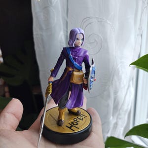May include: A figurine of a character in a purple outfit, holding a sword and shield. The figure has short purple hair and is standing on a base that says "HOPE!!" in gold lettering. The figure is approximately 4 inches tall.