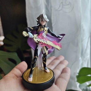 May include: A detailed figurine of a character with silver hair, wearing a black and gold outfit with a purple cape. The figure is holding a pink and gold sword. The base is black with a gold emblem.