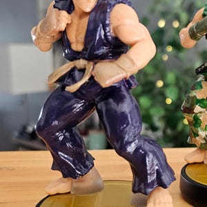 May include: A figurine of Ryu from Street Fighter, posed in a fighting stance. The figure is wearing a dark blue gi, with a red headband and hair. The figure is on a black and gold base.