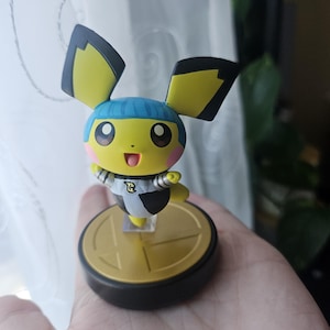 Puede incluir: Figura de Pichu amarillo y negro con gorra azul y mejillas rosas, sobre una base dorada y negra. Figura coleccionable de un personaje de videojuego.