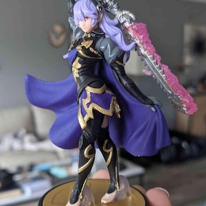 May include: A figurine of a female character with lavender hair, wearing a black and gold outfit with a purple cape. She holds a silver sword with a pink energy blade. The figure stands on a gold base.