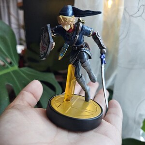 May include: A detailed figurine of a character from The Legend of Zelda series. The figure is in a dynamic pose, holding a sword and shield. The character wears a blue tunic and hat, with blonde hair. The base is black and gold.