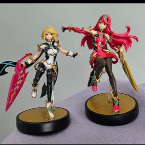 Puede incluir: Dos figuras de acción de personajes femeninos. Una figura tiene cabello rubio, un atuendo blanco, negro y dorado, y una espada rosa y negra. La otra figura tiene cabello rojo, un atuendo rojo y dorado y una espada dorada. Ambas figuras están sobre bases circulares negras y doradas.