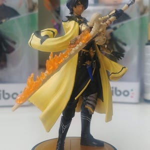 May include: A detailed figurine of a character in a yellow cloak and black outfit, holding a sword with orange flame effects. The figure stands on a black and gold circular base. The background shows other similar figures.
