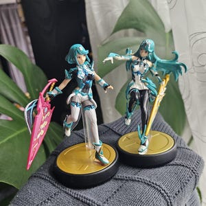 May include: Two anime-style figurines. The figures have blue hair and wear white, black, and blue outfits. One holds a pink and black sword, the other a gold sword. They stand on gold-colored bases with black rims.
