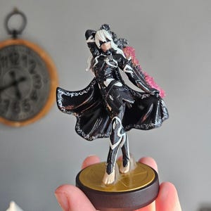 May include: A detailed figurine of a character in a black and white outfit with a flowing cape and white hair. The figure is on a round, gold-colored base. The background is blurred, with a clock visible.