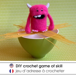 Puede incluir: Un monstruo de crochet rosa en equilibrio sobre fideos de espagueti en un tazón verde. El texto "DIY crochet game of skill" y "jeu d'adresse à crocheter" está debajo de la imagen.