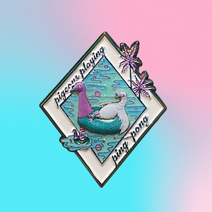 May include: A white enamel pin with a pink and purple pigeon and a white pigeon playing ping pong in a pool. The pin has a blue and green background with palm trees and the text "pigeons playing ping pong".