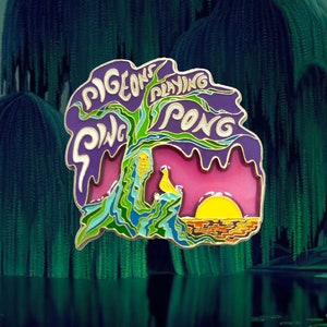 May include: A gold-colored enamel pin with a purple, green, and blue design. The pin features a tree with the words "Pigeons Playing Ping Pong" written around it. The background is pink with a yellow sun.