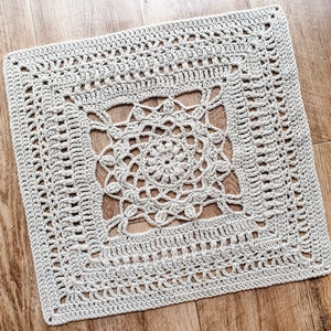 May include: A square crocheted doily with a delicate, intricate design in a light beige color. The doily has a central motif with a circular pattern and a border with a repeating pattern.