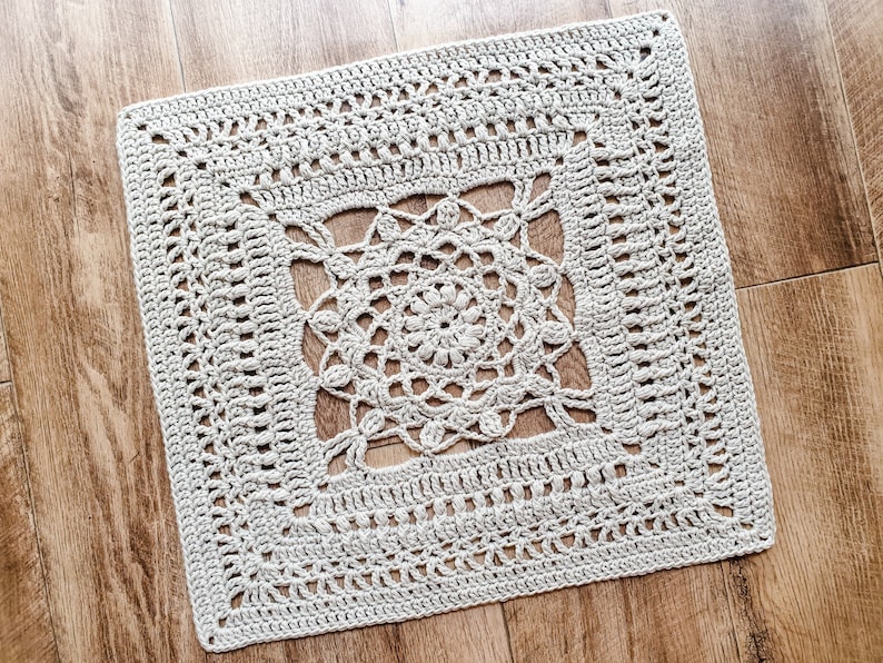 May include: A square crocheted doily with a delicate, intricate design in a light beige colour. The doily has a central motif with a circular pattern and a border with a repeating pattern.
