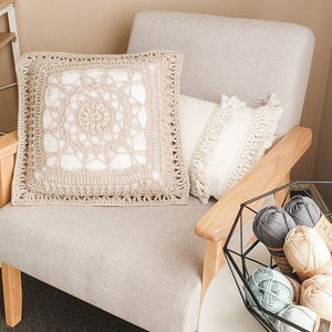 May include: A beige crocheted cushion with a white centre design sits on a light beige armchair with a white cushion with fringe. A black metal bowl filled with colourful yarn balls sits on a white table.