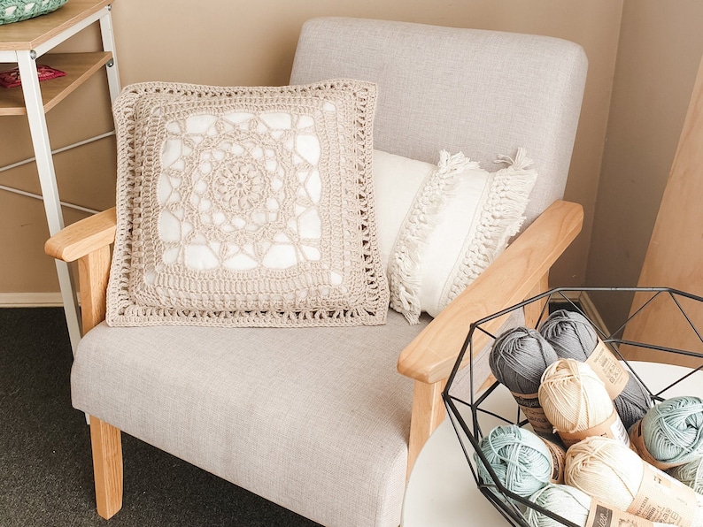 May include: A beige crocheted cushion with a white centre design sits on a light beige armchair with a white cushion with fringe. A black metal bowl filled with colourful yarn balls sits on a white table.