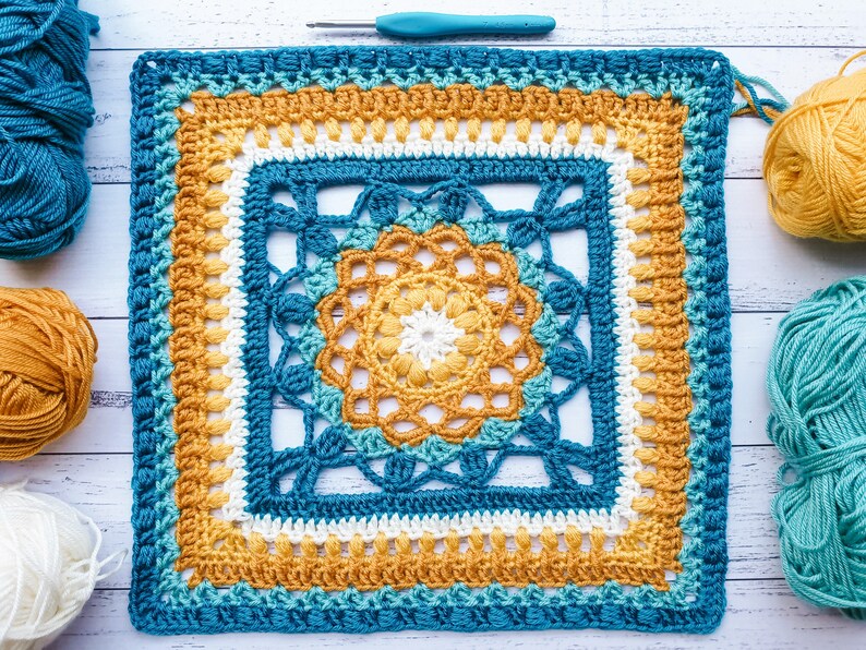 May include: A crocheted square featuring a geometric design in shades of blue, yellow and white. The square is worked using a variety of stitches, including single crochet, double crochet and treble crochet.