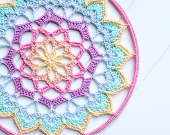Puede incluir: Un mandala de crochet arcoíris con un anillo exterior rosa. El mandala está hecho con una variedad de colores pastel, incluyendo amarillo, azul claro, rosa y morado.