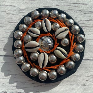 以下が含まれることがあります： 中央に銀色の装飾が施され、オレンジ色の糸と白い貝殻で囲まれた、丸い黒色の装飾品。銀色の金属スタッドが外側の縁を縁取り、ユニークで質感のあるデザインを作り出しています。