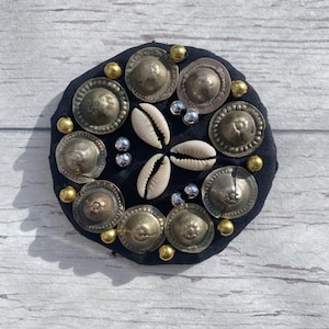 May include: A circular black fabric embellishment with silver metal discs, gold beads, and white seashells. The design is symmetrical, with the seashells forming a central pattern.