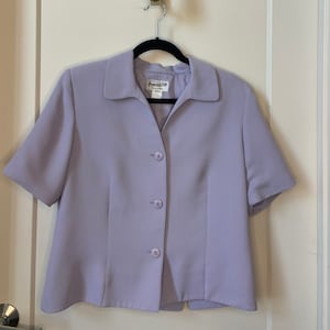 May include: A lavender short-sleeved blazer with a collared neckline and four buttons down the front. The blazer is hanging on a hanger with a pineapple-shaped hook.