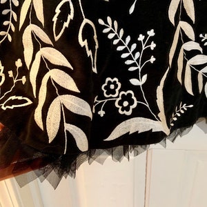 May include: Black tulle skirt with white embroidered floral pattern. The skirt has a scalloped hem.