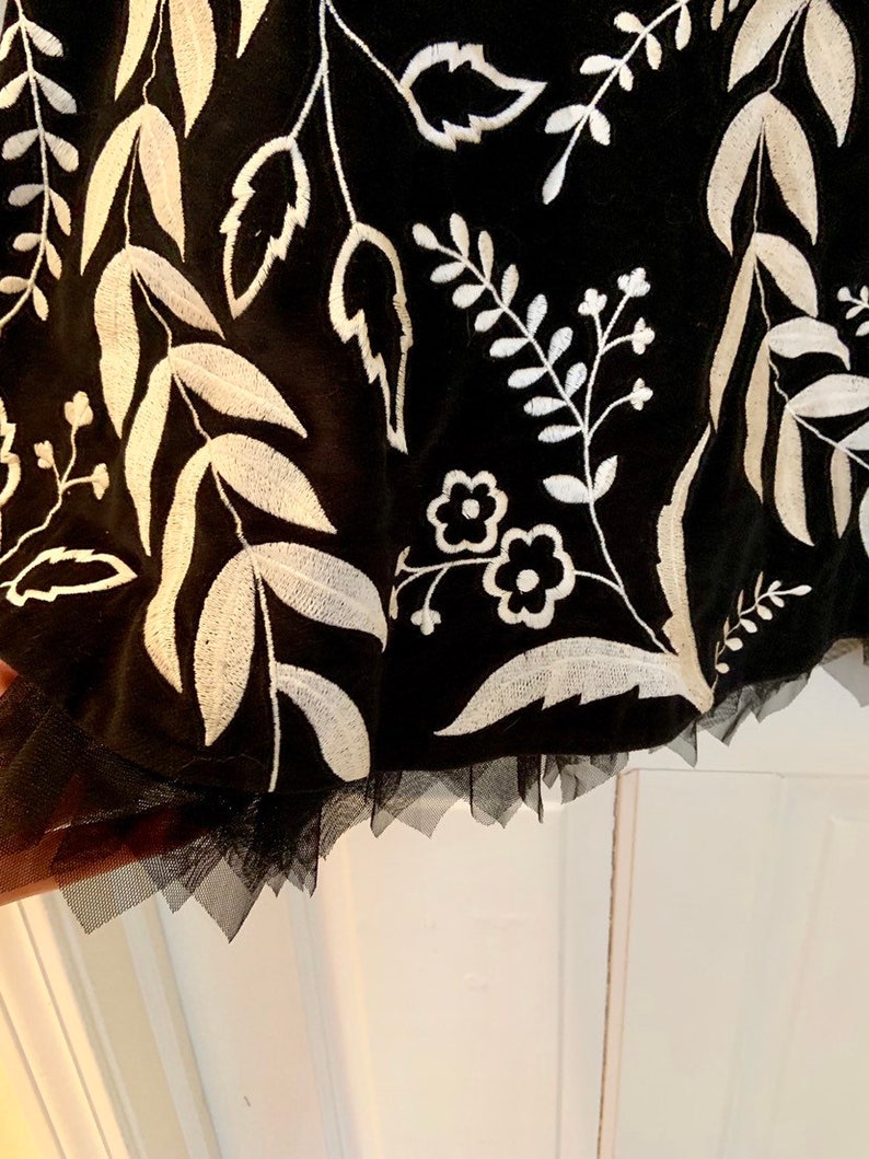 May include: Black tulle skirt with white embroidered floral pattern. The skirt has a scalloped hem.