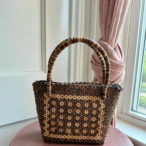 Może przedstawiać: Mała, brązowa torebka z koralikami, zamykana na zamek błyskawiczny i z dwoma zaokrąglonymi uchwytami. Torebka jest ozdobiona okrągłymi koralikami w powtarzającym się wzorze. Torebka stoi na różowej powierzchni.