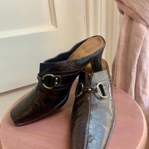 May include: A pair of brown leather heeled mules with gold buckles and studs. The shoes have a textured, crocodile-embossed finish.