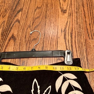 May include: Black suede skirt with white embroidered floral design. The skirt is hanging on a black hanger and has a measuring tape across the top.