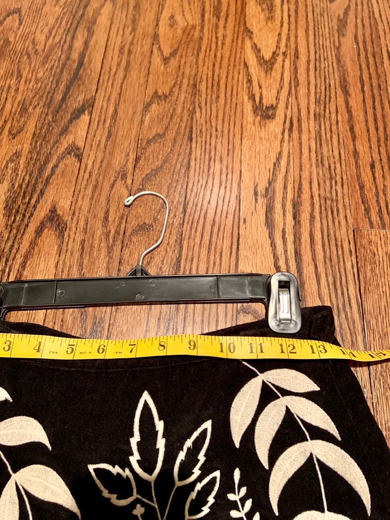 May include: Black suede skirt with white embroidered floral design. The skirt is hanging on a black hanger and has a measuring tape across the top.