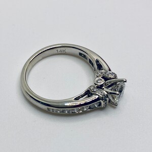 May include: A silver ring with a large, clear, heart-shaped center stone. The band is engraved with "14K" and is adorned with smaller, clear stones. The ring is set against a white background.