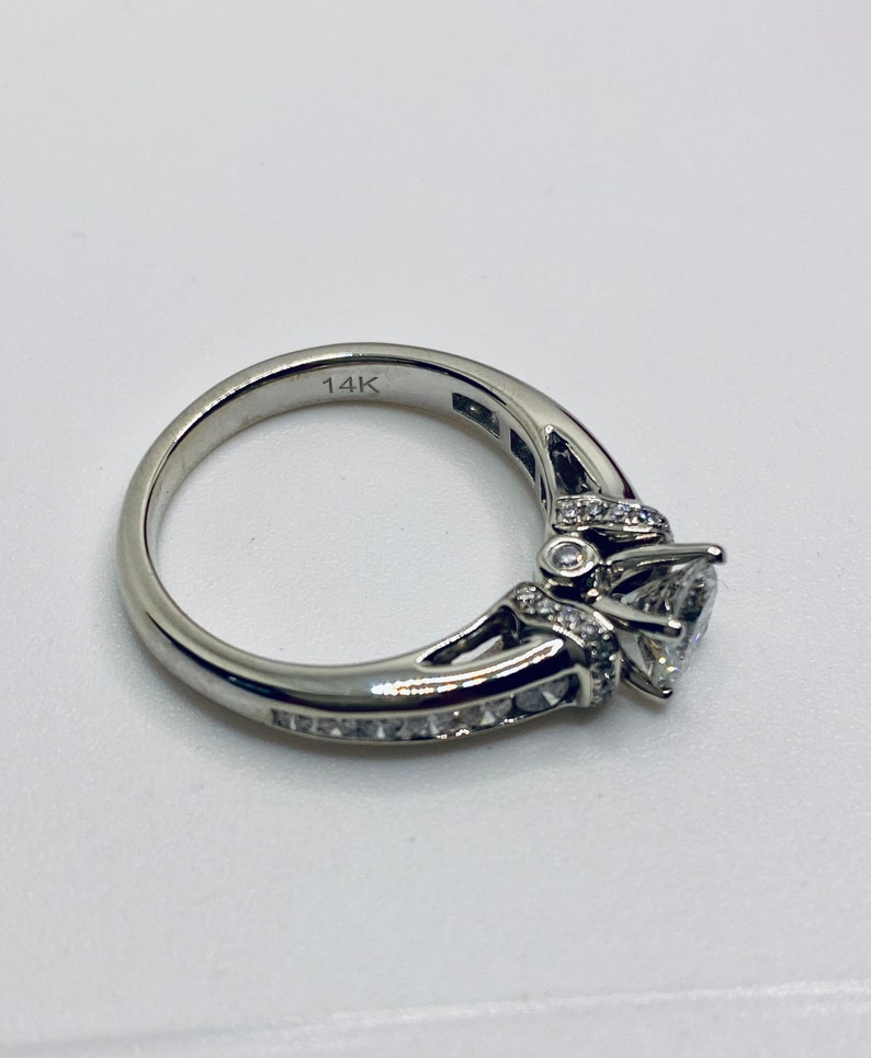 May include: A silver ring with a large, clear, heart-shaped center stone. The band is engraved with "14K" and is adorned with smaller, clear stones. The ring is set against a white background.