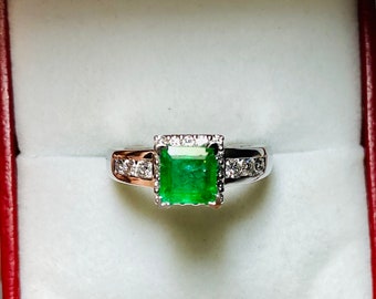 May include: A white gold ring with a large emerald green gemstone surrounded by a halo of smaller diamonds. The ring is set in a white gold band with a row of diamonds on each side.