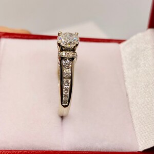 May include: A white gold ring with a large, round, clear diamond center stone. The band features smaller diamonds set along the sides. The ring is displayed inside a red jewelry box with a white satin lining.