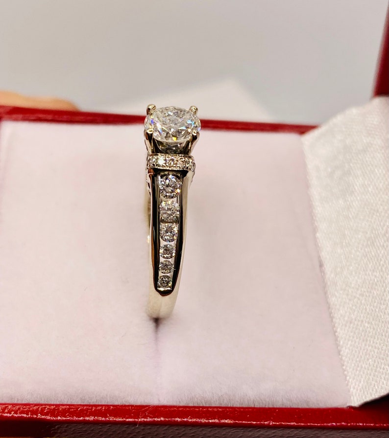 May include: A white gold ring with a large, round, clear diamond center stone. The band features smaller diamonds set along the sides. The ring is displayed inside a red jewelry box with a white satin lining.