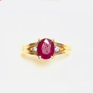 Könnte beinhalten: Ein goldener Ring mit einem großen ovalen Rubin und zwei kleineren Diamanten.