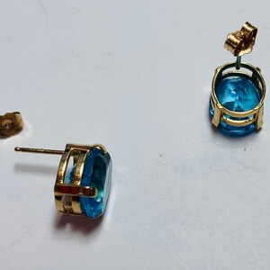 May include: A pair of gold earrings with blue topaz gemstones. The earrings have a simple design with a round gemstone set in a gold setting.