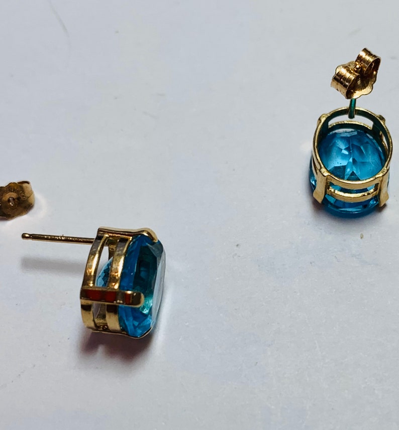 May include: A pair of gold earrings with blue topaz gemstones. The earrings have a simple design with a round gemstone set in a gold setting.