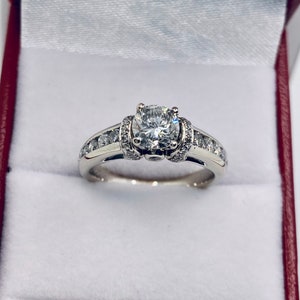May include: A diamond engagement ring with a large, round center stone and smaller diamonds set along the band. The ring is displayed inside a red and white jewelry box, showcasing its brilliance and design.