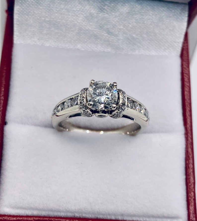 May include: A diamond engagement ring with a large, round center stone and smaller diamonds set along the band. The ring is displayed inside a red and white jewelry box, showcasing its brilliance and design.
