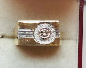 May include: A gold and silver ring with a large, round diamond in the center. The ring is set in a rectangular band with a textured surface.