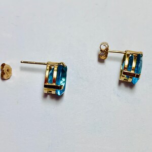 May include: A pair of gold earrings with blue topaz gemstones. The earrings have a simple design with a single stone set in a gold setting.