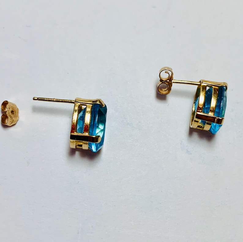 May include: A pair of gold earrings with blue topaz gemstones. The earrings have a simple design with a single stone set in a gold setting.