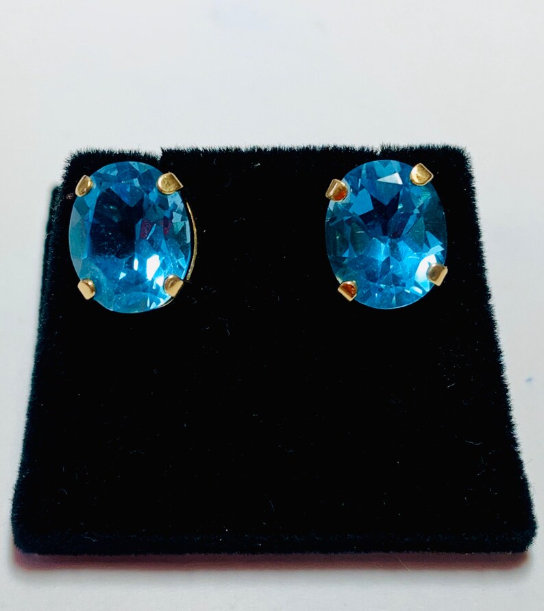 May include: A pair of oval-shaped blue topaz earrings with gold settings. The earrings are displayed on a black velvet background.